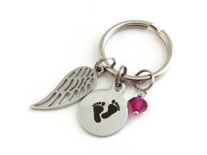 a single angel wing charm, a laser engraved baby footprints charm and a red crystal charm on a keyring