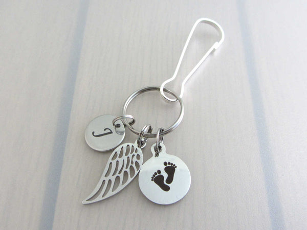 stainless steel laser engraved capital initial letter disc charm, a single angel wing and a laser engraved baby footprints charm on a bag charm with snap clip hook