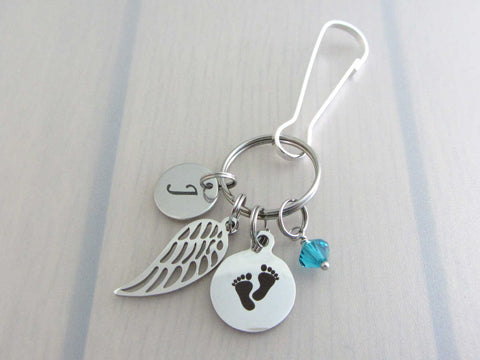 stainless steel laser engraved capital initial letter disc charm, a single angel wing charm, a laser engraved baby footprints charm and a blue/green coloured crystal charm on a bag charm with snap clip hook