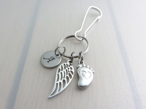 stainless steel laser engraved capital initial letter disc charm, a single angel wing and a single foot charm with indented heart on a bag charm with snap clip hook